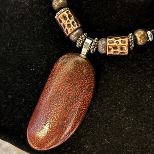 May include: A necklace with a large, reddish-brown pendant and a brown leather cord. The pendant has a textured surface. The necklace also features copper-colored cylindrical beads with a patterned design and smaller beads.