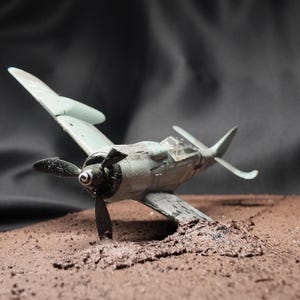 Puede incluir: Maqueta de un avión de combate de la Segunda Guerra Mundial, pintada en tonos grises y verdes, con una hélice negra. El modelo está parcialmente enterrado en una superficie marrón texturizada, sugiriendo un aterrizaje forzoso. El fondo es oscuro.