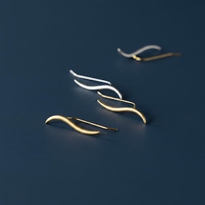 May include: Four minimalist ear climbers in gold and silver tones are arranged on a dark blue surface. The earrings have a curved, wave-like design, with a straight post for insertion into the ear. The earrings are a simple, elegant jewelry design.
