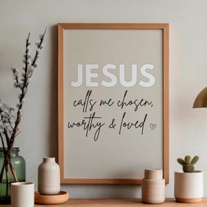 May include: A framed print with the words "JESUS calls me chosen, worthy & loved" in a modern font. The print is displayed with decorative items including a green glass vase, small ceramic vases, and a potted cactus.