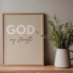 Może przedstawiać: Oprawiony wydruk z napisem "GOD is my strength" w neutralnej palecie kolorów. Grafika jest eksponowana na drewnianej powierzchni, z białym dzbanem z zielenią po prawej stronie.