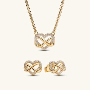 Puede incluir: Un conjunto de collar y pendientes a juego con un diseño de colgante de corazón infinito. Cada pieza está elaborada en metal dorado e incrustada con pequeñas piedras transparentes. El nombre de la marca AURELLE JEWELS aparece arriba a la izquierda.