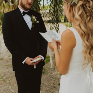 Op de afbeelding: Een zwart smokingjasje met een wit overhemd, zwarte vlinderdas en een kleine bloemencorsage, gecombineerd met een witte mouwloze jurk met een gestrikt detail op de rug. De personen houden witte papieren geloftekaarten met zwarte tekst vast.