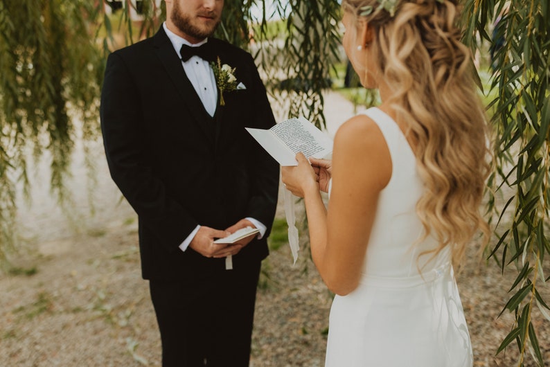 Op de afbeelding: Een zwart smokingjasje met een wit overhemd, zwarte vlinderdas en een kleine bloemencorsage, gecombineerd met een witte mouwloze jurk met een gestrikt detail op de rug. De personen houden witte papieren geloftekaarten met zwarte tekst vast.