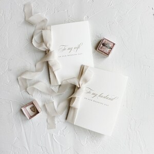 Puede incluir: Dos tarjetas blancas con caligraf&iacute;a que dice "To my wife on our wedding day" y "To my husband on our wedding day". Las tarjetas est&aacute;n atadas con una cinta blanca. Una peque&ntilde;a caja de terciopelo rosa con un anillo dentro est&aacute; sobre la mesa.