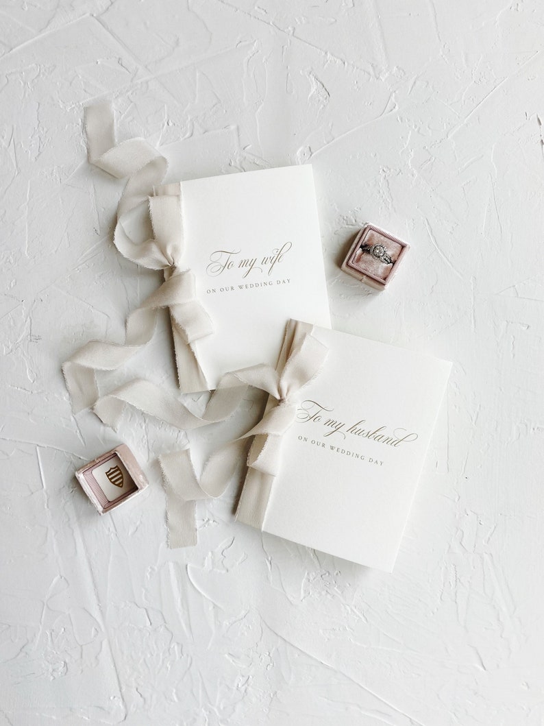 Puede incluir: Dos tarjetas blancas con caligraf&iacute;a que dice "To my wife on our wedding day" y "To my husband on our wedding day". Las tarjetas est&aacute;n atadas con una cinta blanca. Una peque&ntilde;a caja de terciopelo rosa con un anillo dentro est&aacute; sobre la mesa.