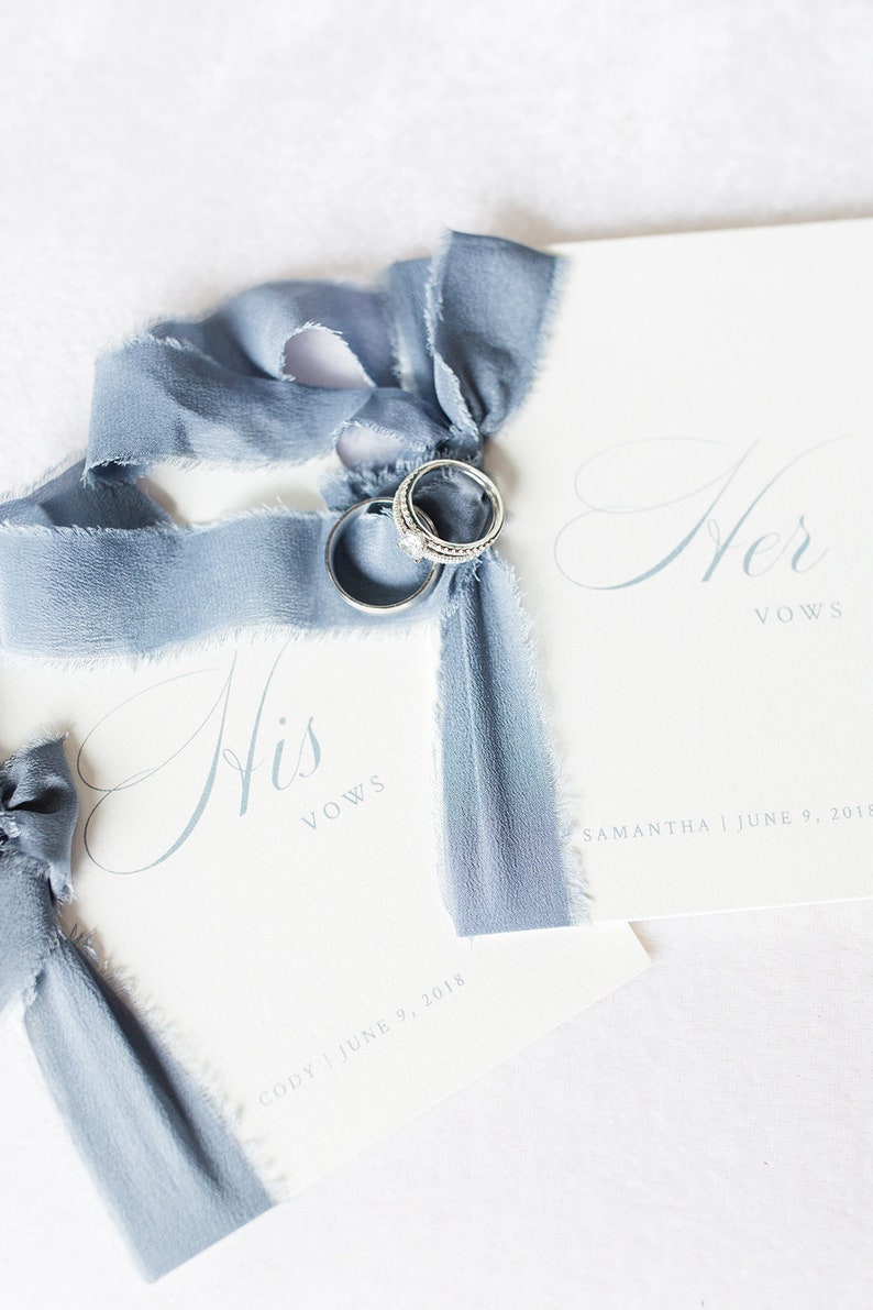 Op de afbeelding: Twee witte gelofteboekjes samengebonden met een blauw, gerafeld zijden lint. E&eacute;n boekje bevat de tekst His Vows met de naam Cody en datum 9 juni 2018. Het andere bevat Her Vows met de naam Samantha en datum 9 juni 2018. Twee zilveren ringen liggen op het lint.