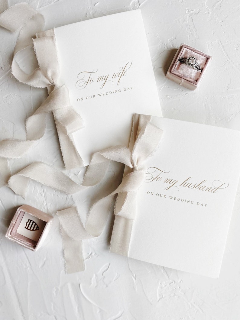 Puede incluir: Dos tarjetas blancas con letras doradas atadas con una cinta beige. Las tarjetas dicen "Para mi esposa en nuestro d&iacute;a de boda" y "Para mi marido en nuestro d&iacute;a de boda".