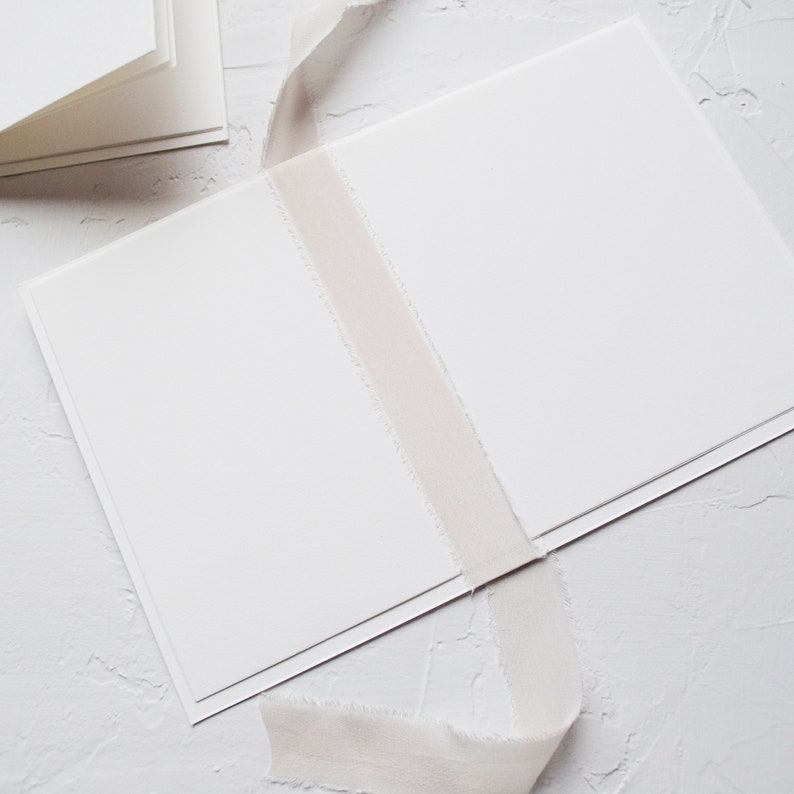 Op de afbeelding: Een open witte kaart of boekje met een glad, mat oppervlak. Een lang, lichtbeige stoffen lint met gerafelde, rauwe randen is verticaal over het midden van de open pagina's gedrapeerd en steekt uit boven de boven- en onderrand van de kaart.