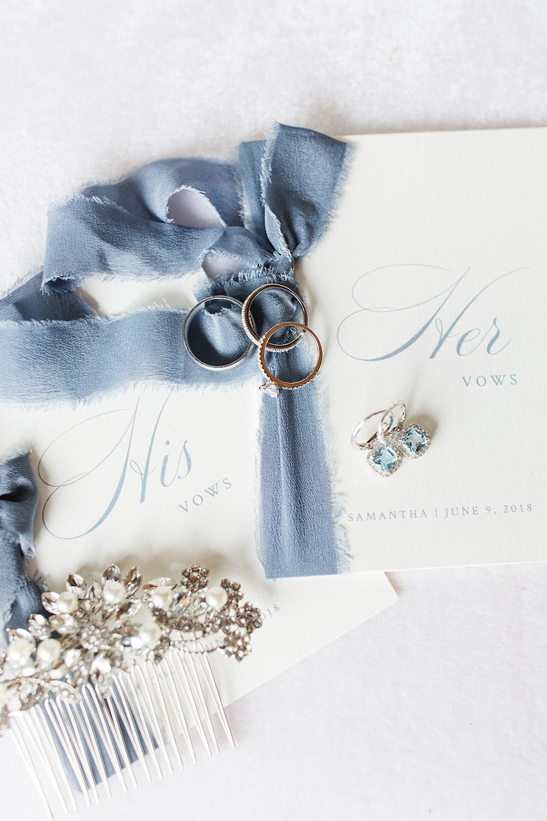 Op de afbeelding: Twee witte gelofteboekjes met de tekst His Vows en Her Vows, blauw zijden lint, drie zilveren ringen, een paar lichtblauwe vierkante oorbellen en een zilveren haarkam met heldere kristallen en witte parels, gedateerd 9 juni 2018 voor Samantha.