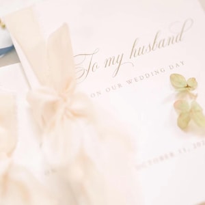 May include: A white card with the gold script text To my husband ON OUR WEDDING DAY and a date at the bottom. The card is tied with a light cream-colored fabric ribbon and decorated with small green floral accents on the side.