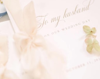 Op de afbeelding: Een witte kaart met de gouden cursieve tekst To my husband ON OUR WEDDING DAY en een datum onderaan. De kaart is vastgebonden met een licht crèmekleurig stoffen lint en versierd met kleine groene florale accenten aan de zijkant.