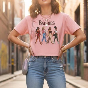 May include: A light pink crop top with the text "THE BADDIES" and a graphic of four women in different outfits. The top is paired with light blue ripped jeans and white sneakers. The person is wearing sunglasses and gold jewelry.