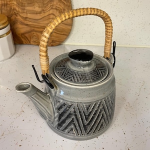 May include: A ceramic teapot with a speckled light gray glaze and a dark gray geometric chevron pattern. It features a black lid knob and a curved handle wrapped in light brown woven rattan, attached to the body with black metal brackets.