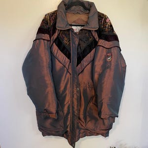 May include: A long-sleeved, iridescent brown and purple puffer jacket featuring a high collar, a front zipper closure with snap buttons, and dark velvet panels on the chest and shoulders. A small rectangular patch with the text Triple F.A.T. Goose is visible.