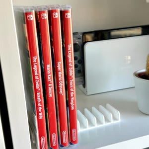 Könnte beinhalten: Ein Regal zeigt vier Nintendo Switch-Spielehüllen mit roten Rücken. Die Spiele sind "The Legend of Zelda: Breath of the Wild", "Mario Kart 8 Deluxe", "Super Mario Odyssey" und "The Legend of Zelda: Tears of the Kingdom". Eine weiße Switch-Konsole und eine Pflanze sind ebenfalls zu sehen.