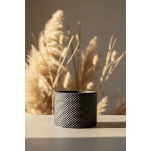 Könnte beinhalten: Ein schwarzer, zylindrischer Pflanzentopf mit einer strukturierten, gepunkteten Oberfläche. Der Topf steht auf einer hellen Oberfläche, mit getrockneten Ziergräsern im Hintergrund. Der Topf ist für den Innen- und Außenbereich geeignet.