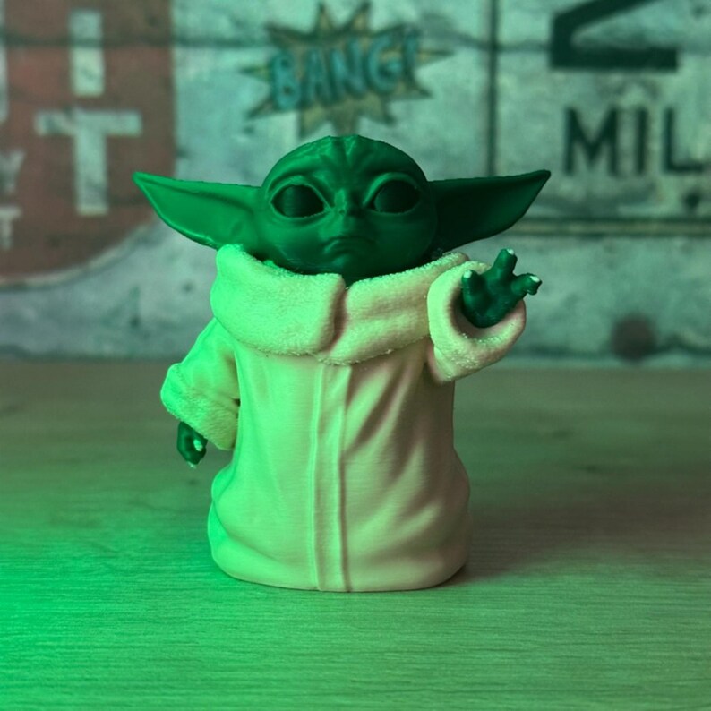 May include: A green figurine of a character with large ears and a cream-colored robe. The figurine is posed with one hand raised. The background features graffiti-style text, including the word "BANG".
