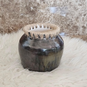 May include: A dark, glazed ceramic vase with a rounded body and a textured, rope-wrapped rim. The vase has a metallic sheen, with a neutral color palette. The interior is white. The vase is sitting on a white, fluffy surface.