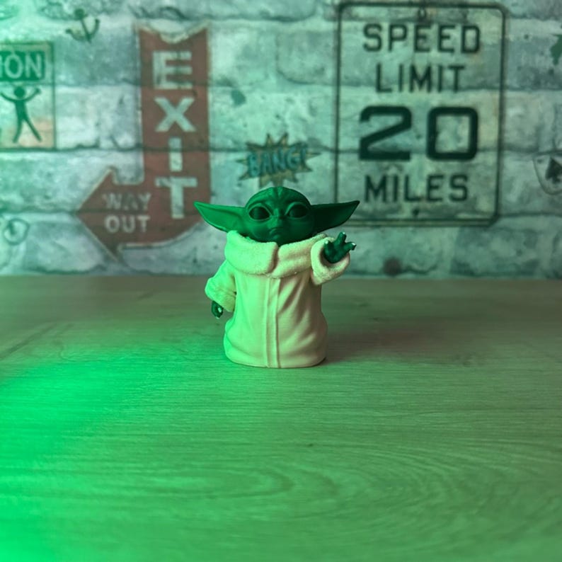 May include: A green figurine of a character with large ears and a tan robe, with one hand raised. The figurine is on a wooden surface, with a brick wall background and a speed limit sign.