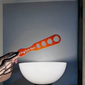 Puede incluir: Una herramienta naranja para medir espaguetis con la inscripción "Spaghetti Mate". La herramienta tiene cuatro agujeros circulares de diferentes tamaños, cada uno etiquetado para una porción diferente. La herramienta se sostiene sobre un cuenco blanco.