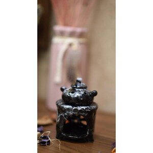 May include: A black and white ceramic oil warmer with a teapot-shaped top. The warmer has a small, open base and a decorative, marbled pattern. The background includes a pink vase with dried flowers.