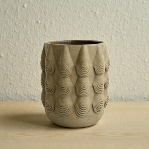 Op de afbeelding: Een beige keramische vaas met een textuur, geometrisch ontwerp. De vaas heeft een herhalend patroon van kegelvormige elementen met gebogen, golfachtige details. De binnenkant van de vaas is donkerder van kleur en staat op een houten oppervlak.