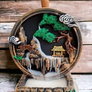 Op de afbeelding: Een decoratieve wierookbrander met een gedetailleerde Aziatische landschapsscène. De ronde, bruin omrande brander heeft een waterval, brug en pagode. Groen gebladerte en een wolkvormig accent dragen bij aan het ontwerp. De basis is een steenachtige structuur.