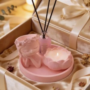 May include: A pink gift set comprising a ceramic figure, a lotus-shaped candle, and a small glass bottle with reeds. The set is presented in a box with a satin ribbon, suitable for home decor or gifting.