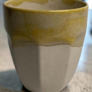 Può includere: Una tazza in ceramica dal design sfaccettato. La parte superiore è di colore giallo caldo, che sfuma in una base bianco sporco macchiettata. La tazza ha un'estetica semplice e moderna, adatta alle bevande.