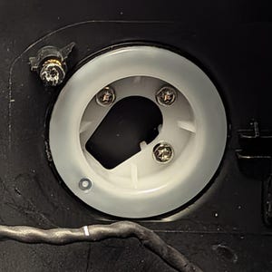 May include: Close-up of a circular, white plastic component with a central opening and three screws. The component is set against a black background, with a small screw and a wire visible.