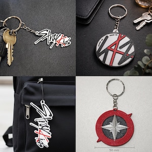 Könnte beinhalten: Eine Sammlung von vier Schlüsselanhängern mit dem Stray Kids-Logo in Weiß, Schwarz und Rot. Ein runder Schlüsselanhänger zeigt ein Kompassdesign mit einer 50 mm Maßangabe. Die Artikel verfügen über Metallringe und Ketten zur Befestigung.
