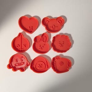 Puede incluir: Un conjunto de ocho cortadores de galletas de plástico rojo con varias caras de personajes y formas, incluyendo un corazón, un koala, un conejo y otras figuras estilizadas con detalles faciales en relieve, dispuestos en cuadrícula.