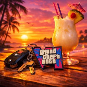 Może przedstawiać: Czarny pilot do samochodu i dwa metalowe klucze przymocowane do breloczka z różowo-niebieskim logo Grand Theft Auto, umieszczone obok szklanki tropikalnego koktajlu z plasterkiem ananasa, czerwoną wisienką i dwiema słomkami.