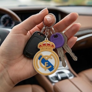 Peut inclure: Un porte-clés avec le logo du Real Madrid, un porte-clés de voiture noir et deux clés. Le porte-clés présente un emblème blanc, bleu et or avec une couronne. Les clés ont des têtes de clés violettes et argentées.