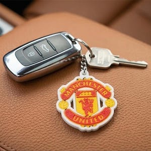 Könnte beinhalten: Ein Autoschlüssel und ein Schlüssel mit einem Manchester United Schlüsselanhänger. Der Schlüsselanhänger ist weiß mit roten und gelben Akzenten und zeigt das Vereinswappen und die Worte "MANCHESTER UNITED". Die Schlüssel und der Schlüsselanhänger liegen auf einer braunen Lederoberfläche.