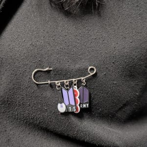 Op de afbeelding: Een zilverkleurige veiligheidsspeld met vier bungelende bedels. De bedels bestaan uit een paars en zwart BTS-logo, een wit en paars cirkelvormig icoon, een verticaal rood-wit gestreept element en een donkerpaarse vijfhoekige vorm met de tekst ARMY.