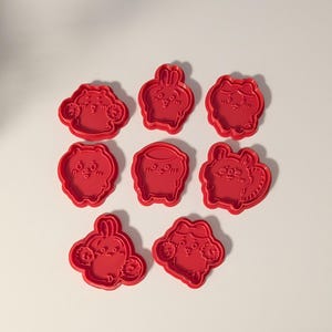 May include: A set of eight red plastic cookie cutters, each featuring a distinct embossed character design with facial features and rounded shapes. The cutters are arranged in a grid pattern, showcasing various cartoon-like figures with expressions.