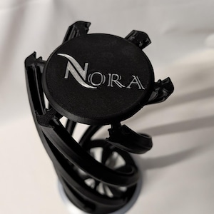 Könnte beinhalten: Ein schwarzes, 3D-gedrucktes Dekorationsobjekt mit rundem Oberteil und spiralförmigem Sockel. Auf der Oberseite steht der Name "Nora" in silberner, stilisierter Schrift. Das Objekt steht vor einem weißen Hintergrund.