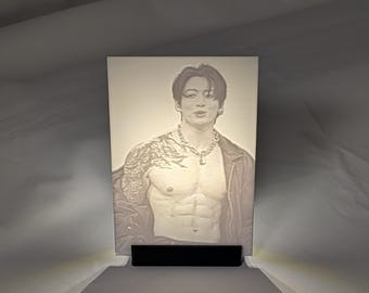 May include: A rectangular, illuminated lithophane panel featuring a grayscale portrait of a person with a muscular torso, wearing a chain necklace and an open jacket. The image is displayed on a small, black, rectangular stand.