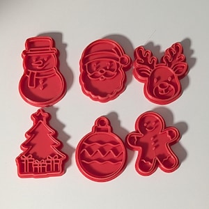 May include: A set of six red plastic cookie cutters featuring holiday shapes including a snowman, Santa Claus, a reindeer, a Christmas tree with presents, a round ornament, and a gingerbread person, all arranged in two rows of three items.
