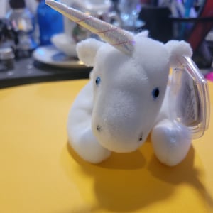 May include: A white plush unicorn toy with a sparkly horn and blue eyes. The unicorn has a tag attached. The toy is on a yellow surface.