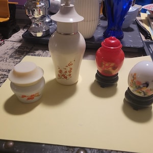 Op de afbeelding: Een verzameling decoratieve keramische items, waaronder een witte pot met deksel, een witte fles met rode bloemmotieven, een rode pot met deksel en een wit eivormig ornament met een vogel- en bloemdesign. Elk item staat op een standaard.