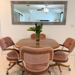 May include: A dining set featuring a rounded wooden table and four swivel chairs upholstered in dusty pink fabric with wooden frames and metal casters. A clear glass vase with green foliage sits on the table, below a large rectangular wall mirror.