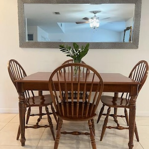 May include: A wooden dining table with four matching chairs. The chairs have a spindle back design. A large rectangular mirror hangs on the wall above the table. A vase of greenery sits on the table.