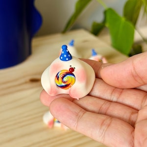 May include: A whimsical, handmade figurine of a smiling, white creature with pink cheeks, a blue party hat, and a colorful lollipop. The figurine has a red nose and is held in a hand. The background is blurred.