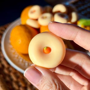 May include: A small, round, pale yellow confection with a smooth surface and a central indentation featuring a tiny, textured orange accent. The treat is held between fingers, with a plate of similar items visible in the background.