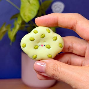 May include: A pale yellow, cloud-shaped object featuring a smooth surface decorated with small, raised olive green leaf-like patterns. The center of the object displays a simple face with two tiny dots for eyes and a small, bright pink dot for a mouth.