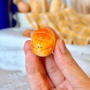 以下が含まれることがあります： 表面に笑顔が彫られた、オレンジと黄色のミニチュアパン。小さな焼き菓子は、手で持たれており、質感が良く、詳細な外観をしています。背景には、ぼやけたパンと黄色のチェック柄の布が含まれています。