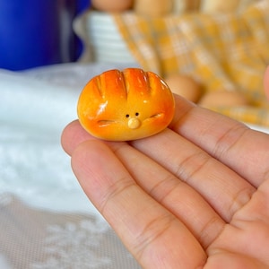 May include: A miniature, orange-yellow bread roll with a cartoon face. The small, round loaf has a textured surface with vertical lines and a small, round face. The background includes more bread and eggs, suggesting a bakery theme.